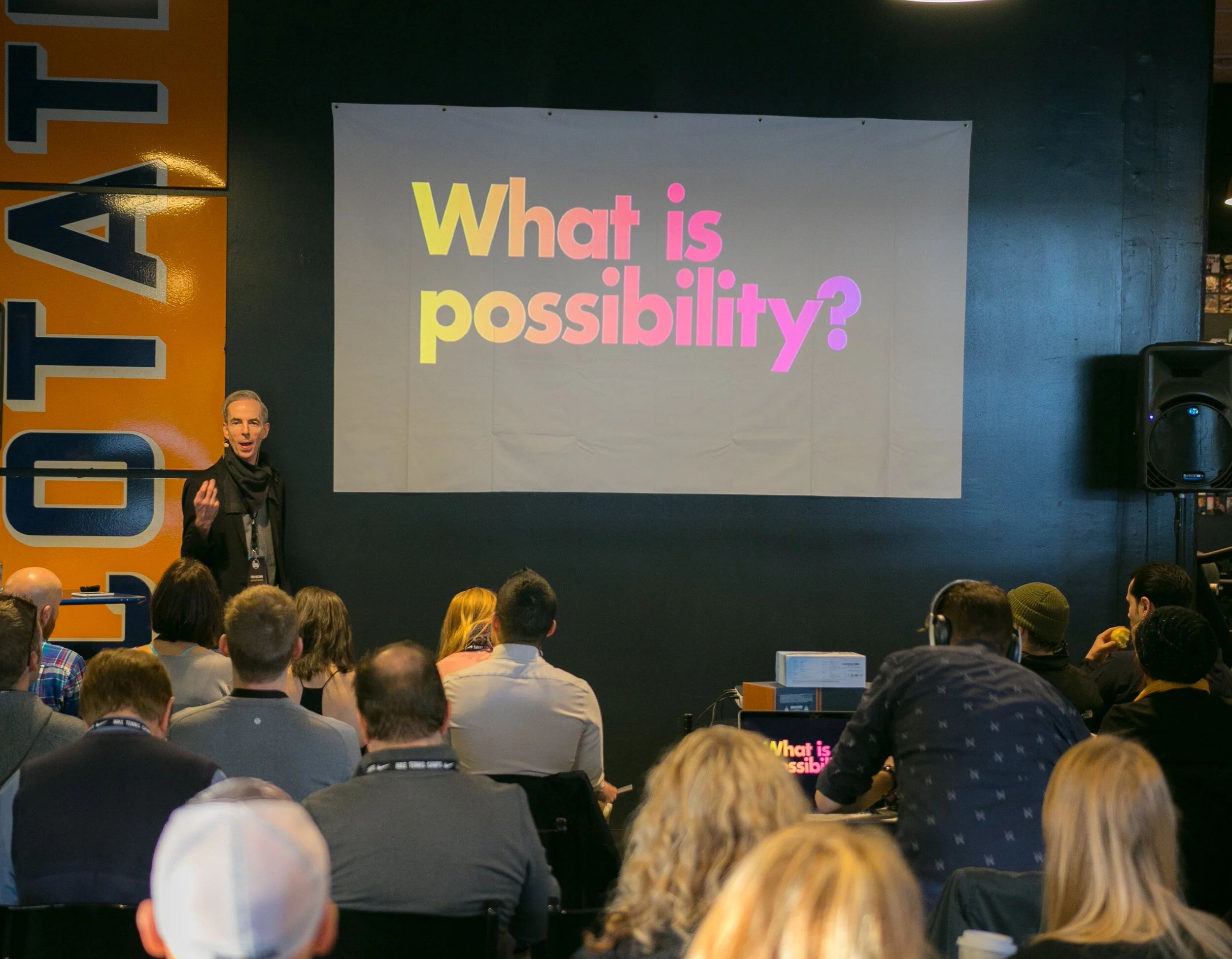 Presenter on stage with a 'What is possibility?' projection behind him at a Business Movement event