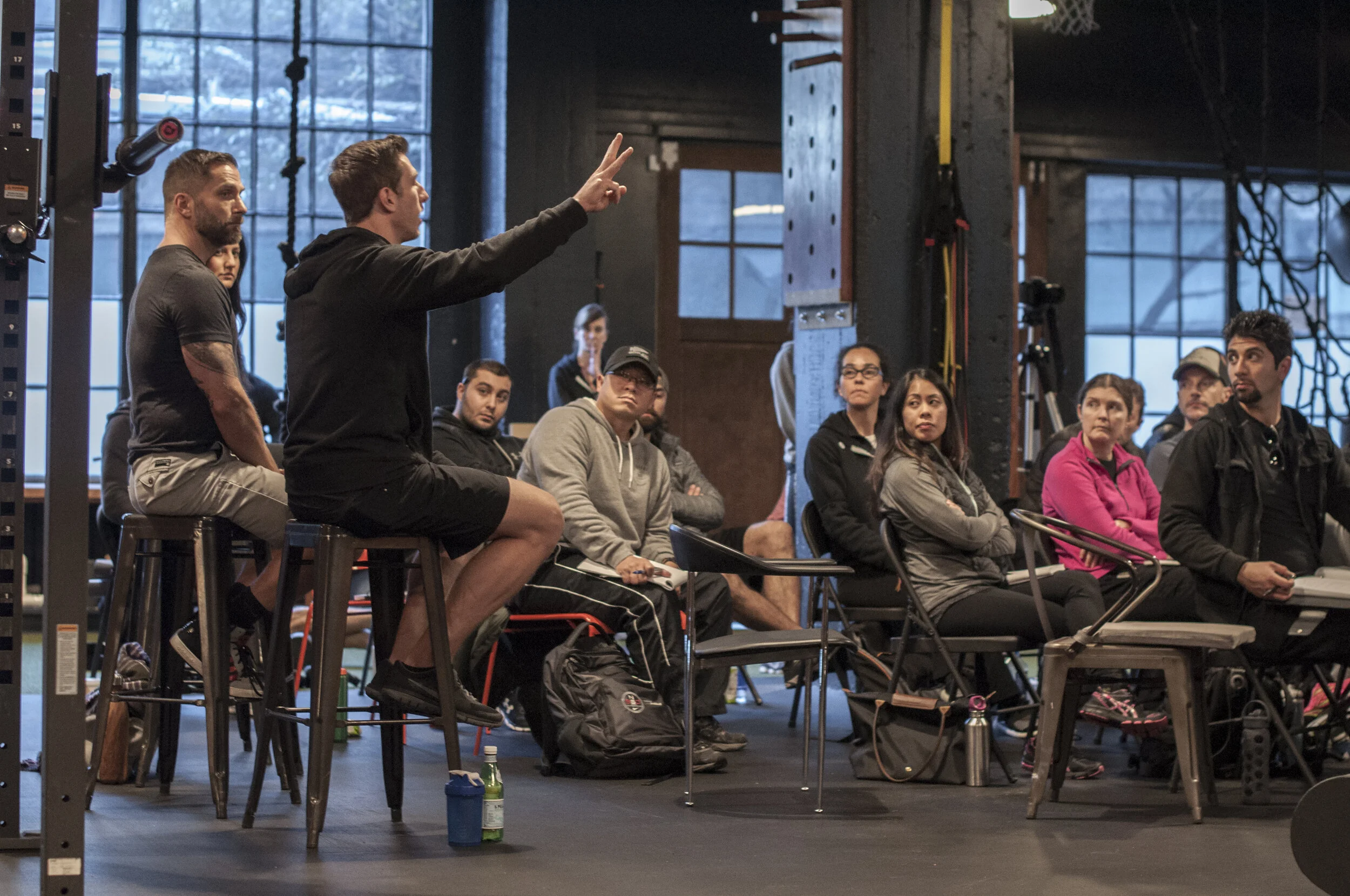 Billy Polson leading a fitness business coaching session in a gym, seated on a barstool gesturing to an attentive audience