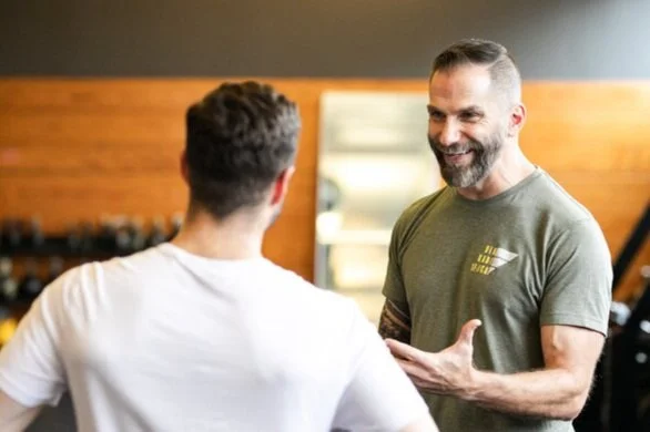 A grey-bearded coach in an olive Diakadi shirt talking with a client during a coaching session in a gym