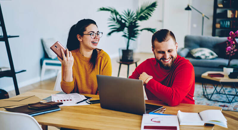 A woman in a yellow sweater and a man in a red sweater laughing together at a laptop