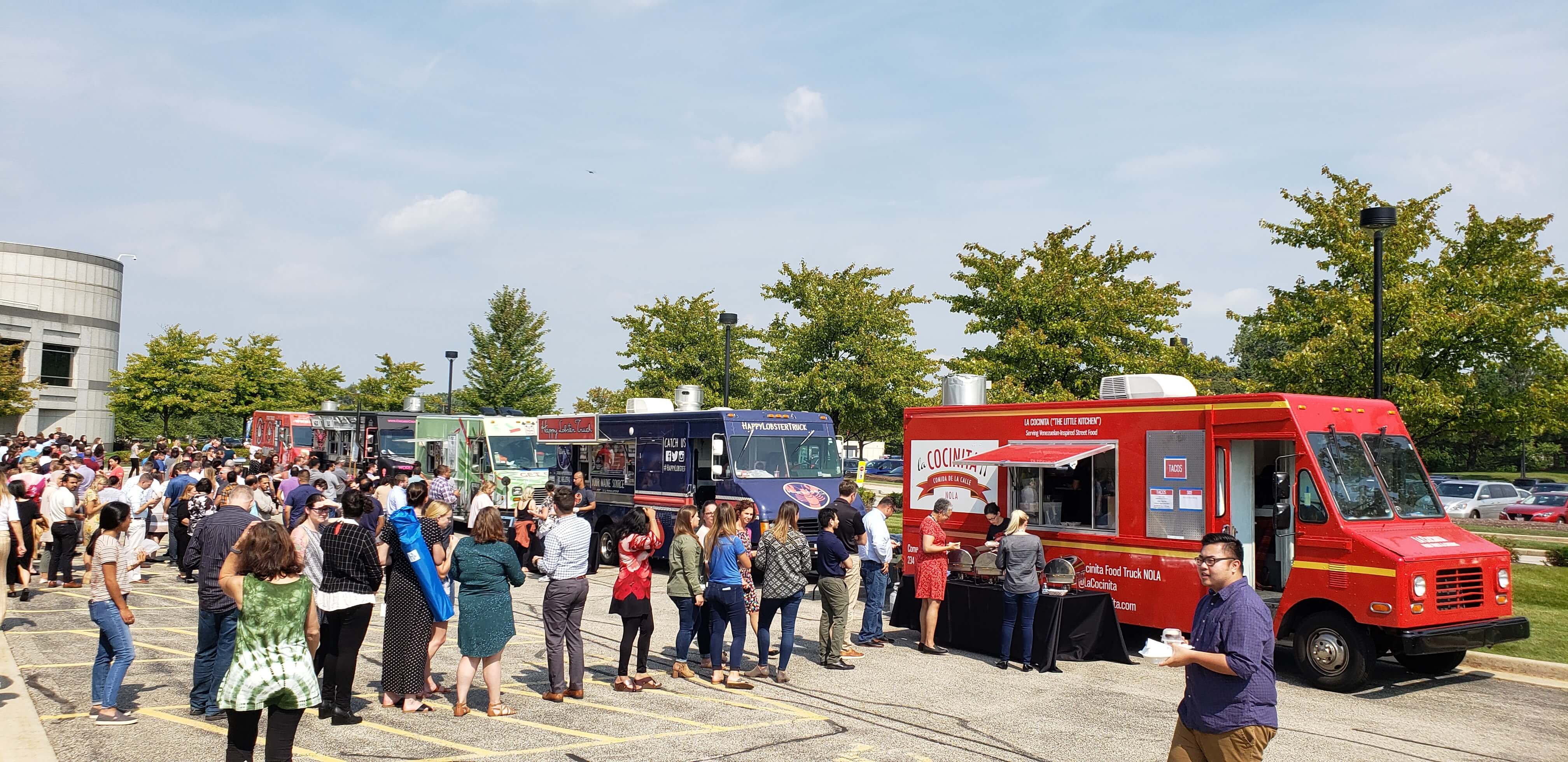 Large crowd lined up at multiple food trucks in a suburban parking lot for a corporate catering event