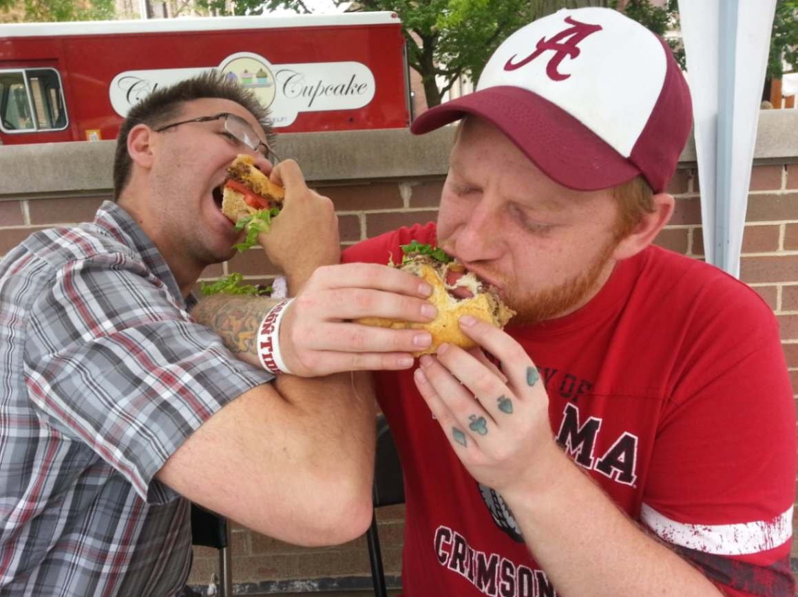 Two men enthusiastically eating large sandwiches at an outdoor food truck event