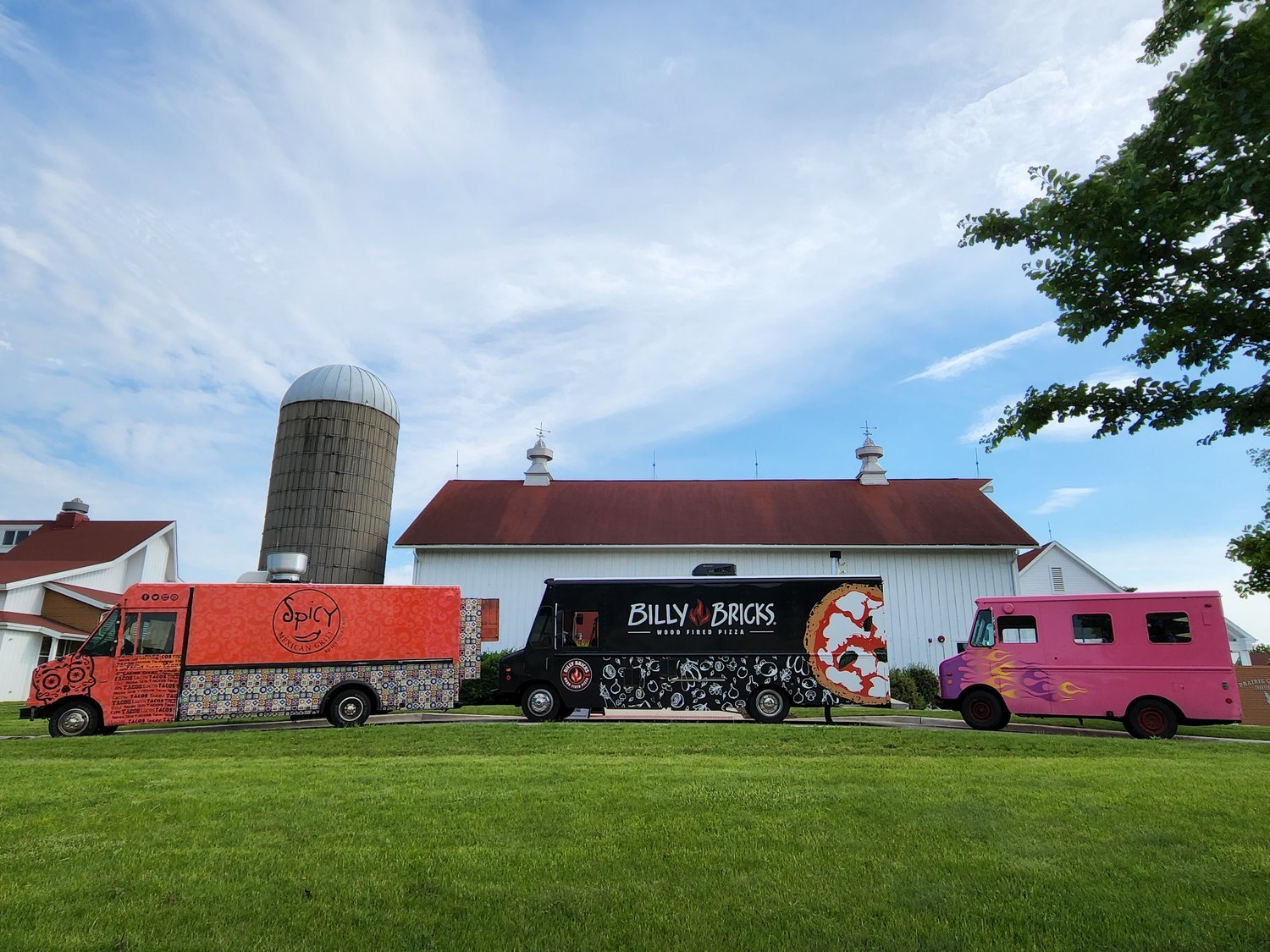 Three colorful food trucks parked in front of a white barn with silo