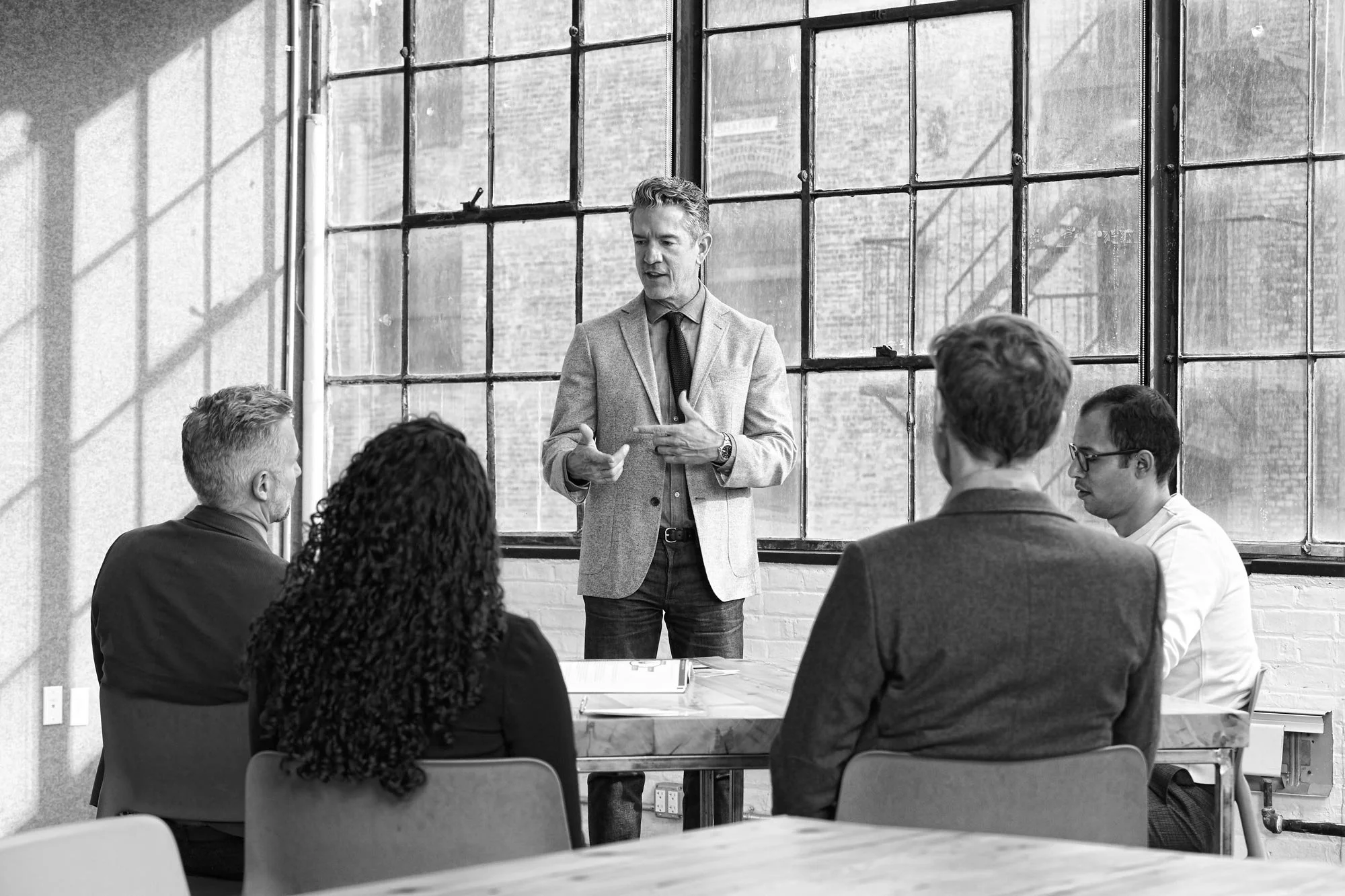 A facilitator in a blazer leading a workshop for seated attendees in a sunlit New York loft