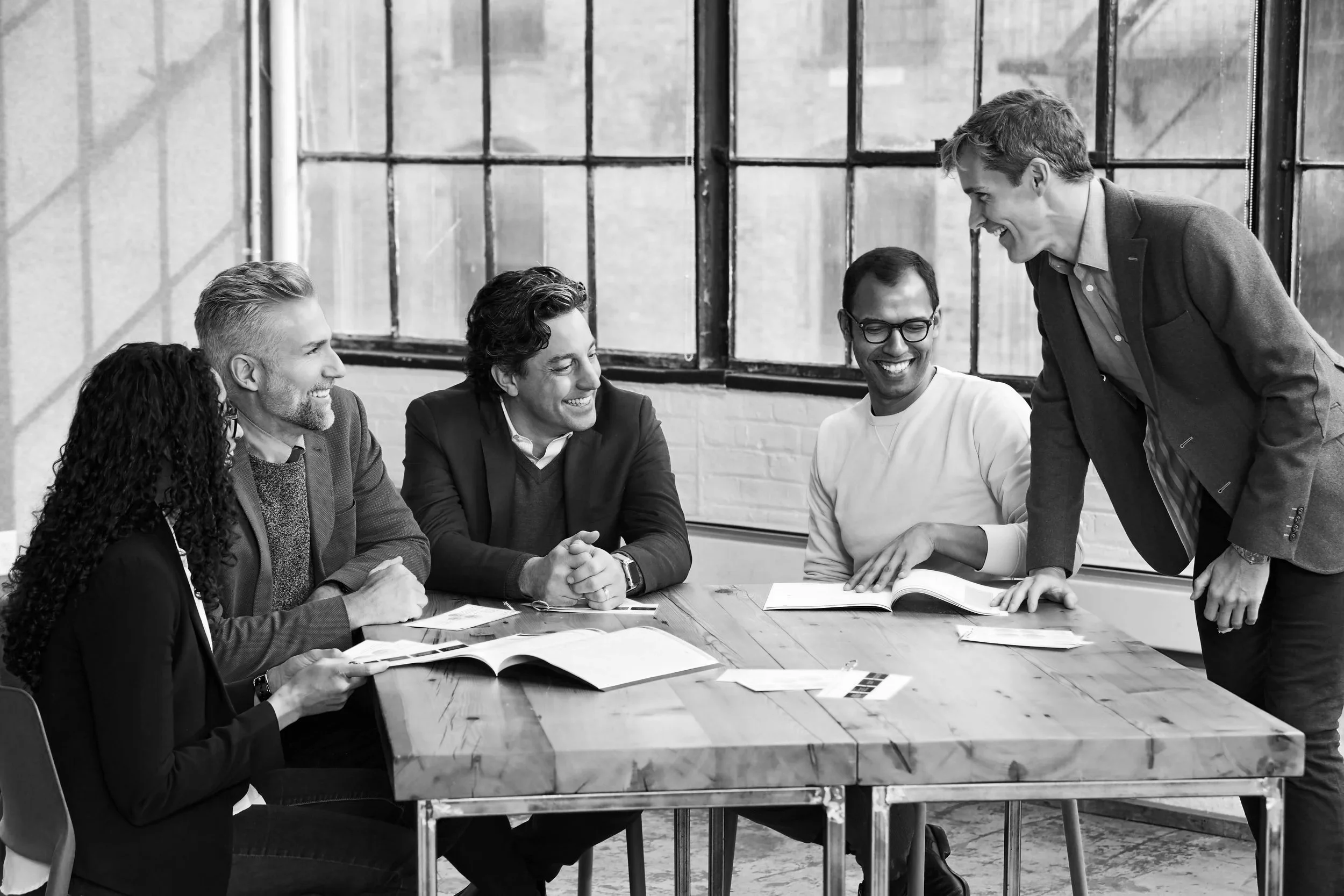 Five diverse professionals laughing and collaborating around a table in an industrial New York loft