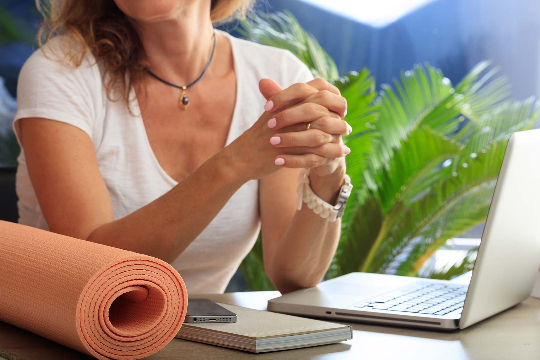 Woman seated at a desk with a rolled yoga mat, laptop, phone and notebook during a virtual wellness session