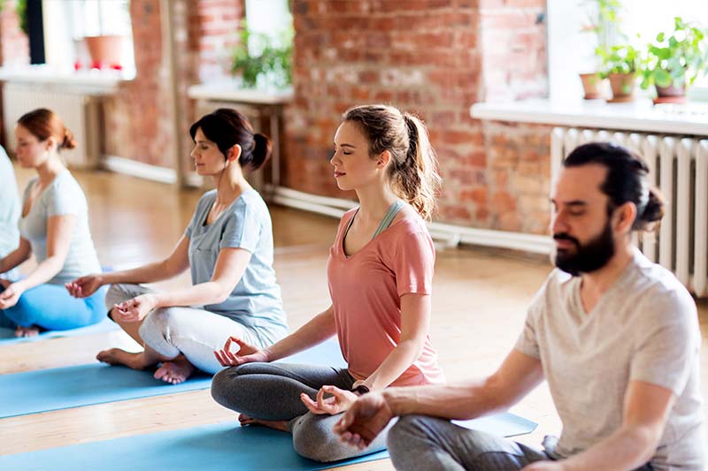 Four adults meditating in lotus pose on yoga mats in a brick-walled studio