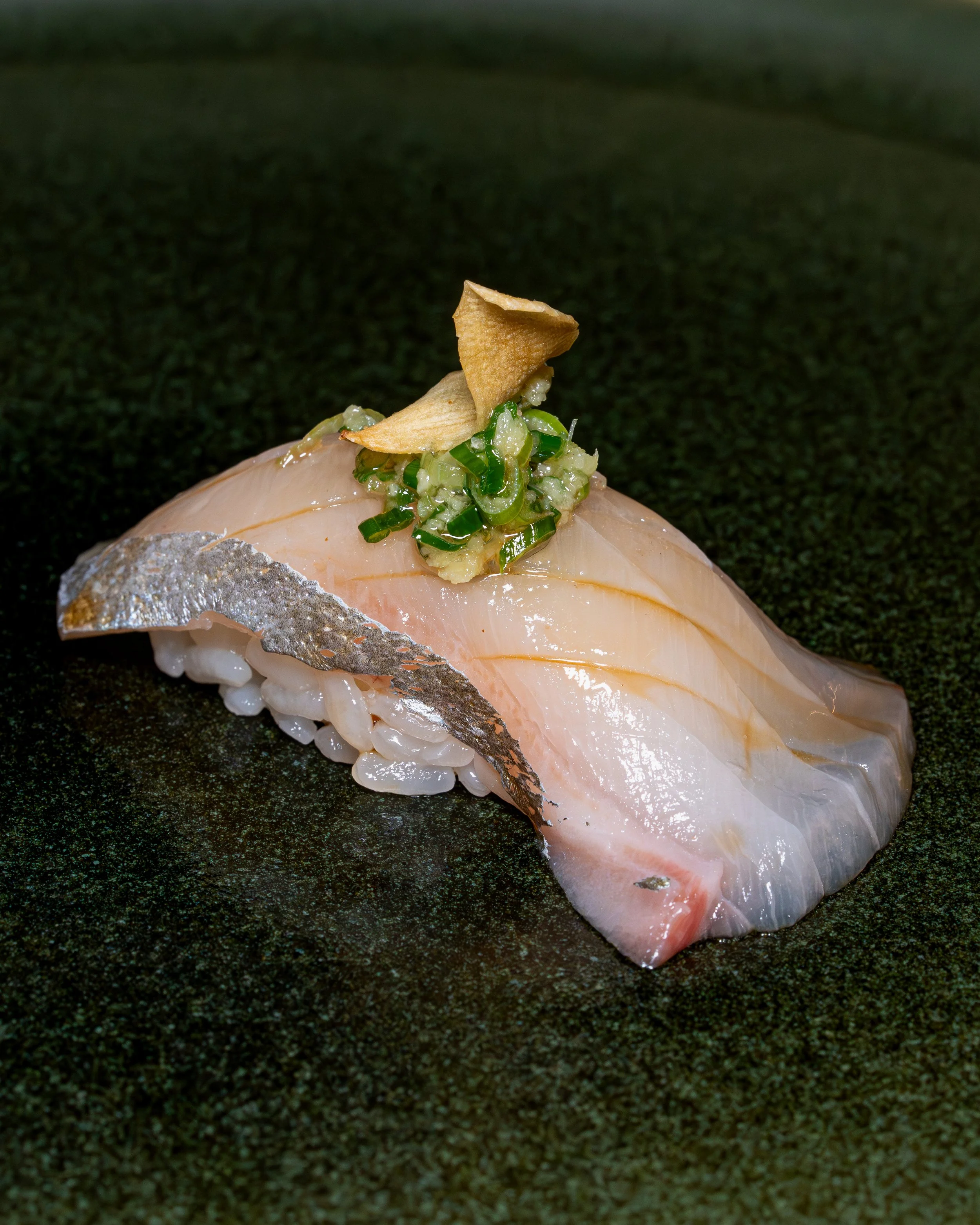 Shima aji nigiri with garlic chip and ginger scallion on dark stone
