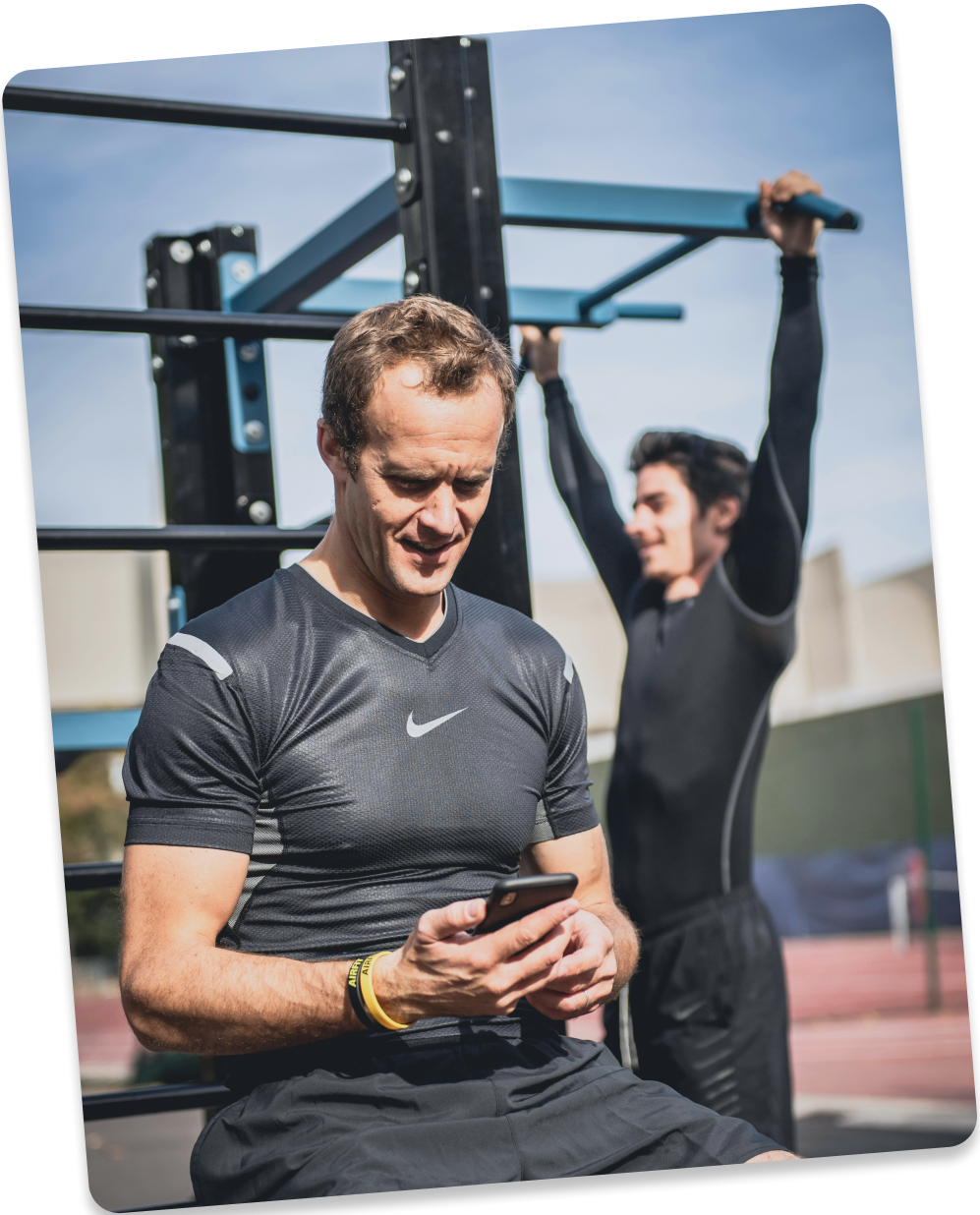 Man checking his phone at an outdoor gym while another man does pull-ups behind him