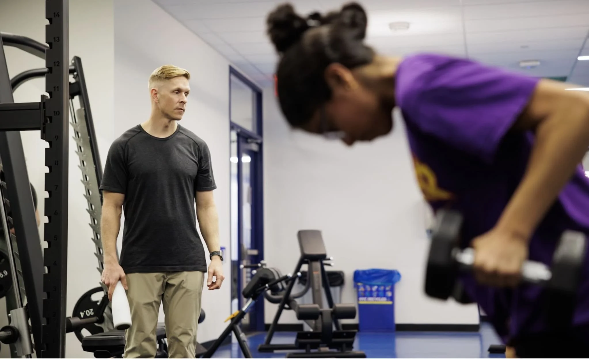 Dan Daly in a dark tee coaching a female athlete through a dumbbell row in the gym.