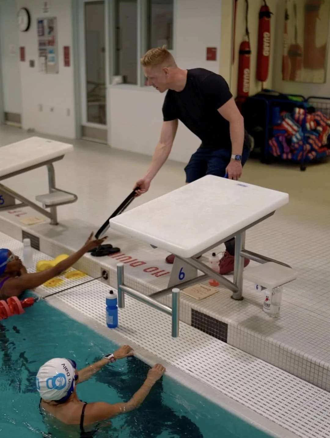 Coach Dan Daly in a black shirt leaning over the pool edge instructing a swimmer in the lane.