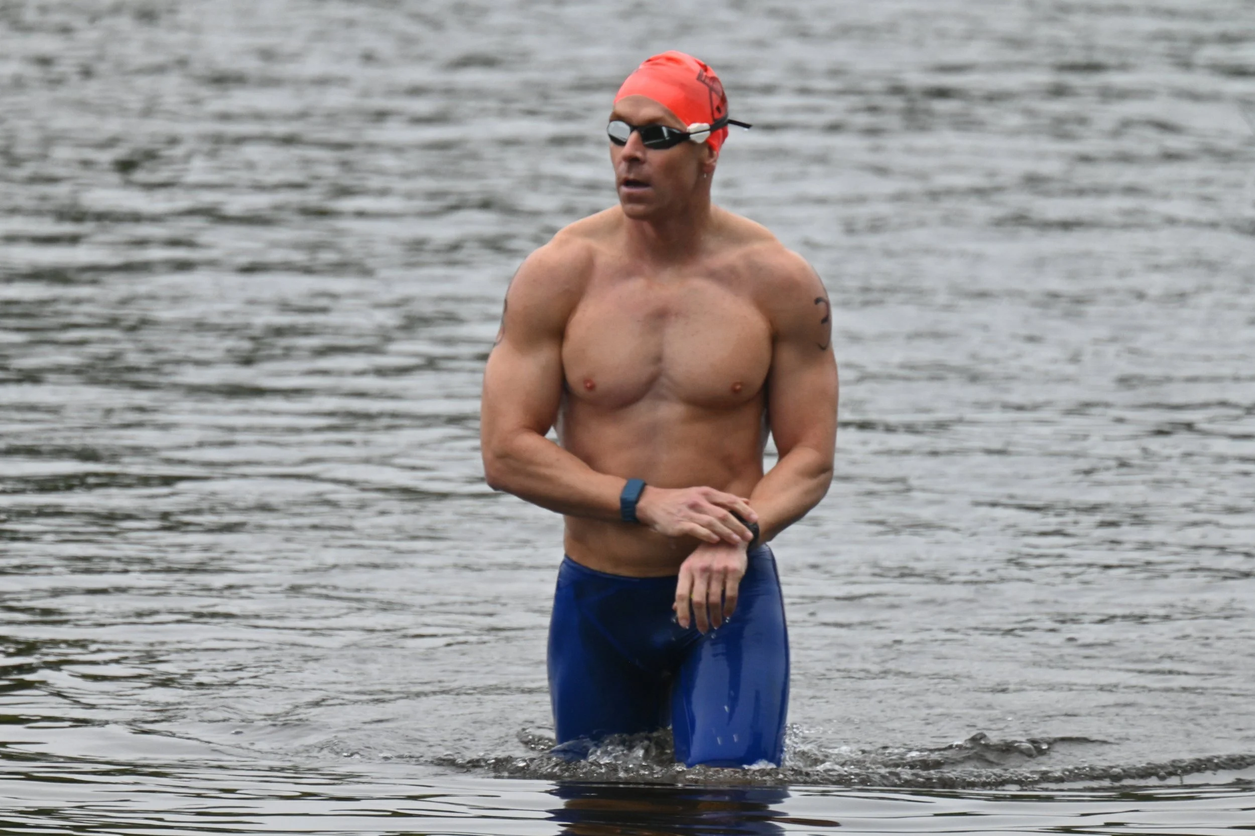 Dan Daly in an orange swim cap and goggles standing waist-deep in open water.