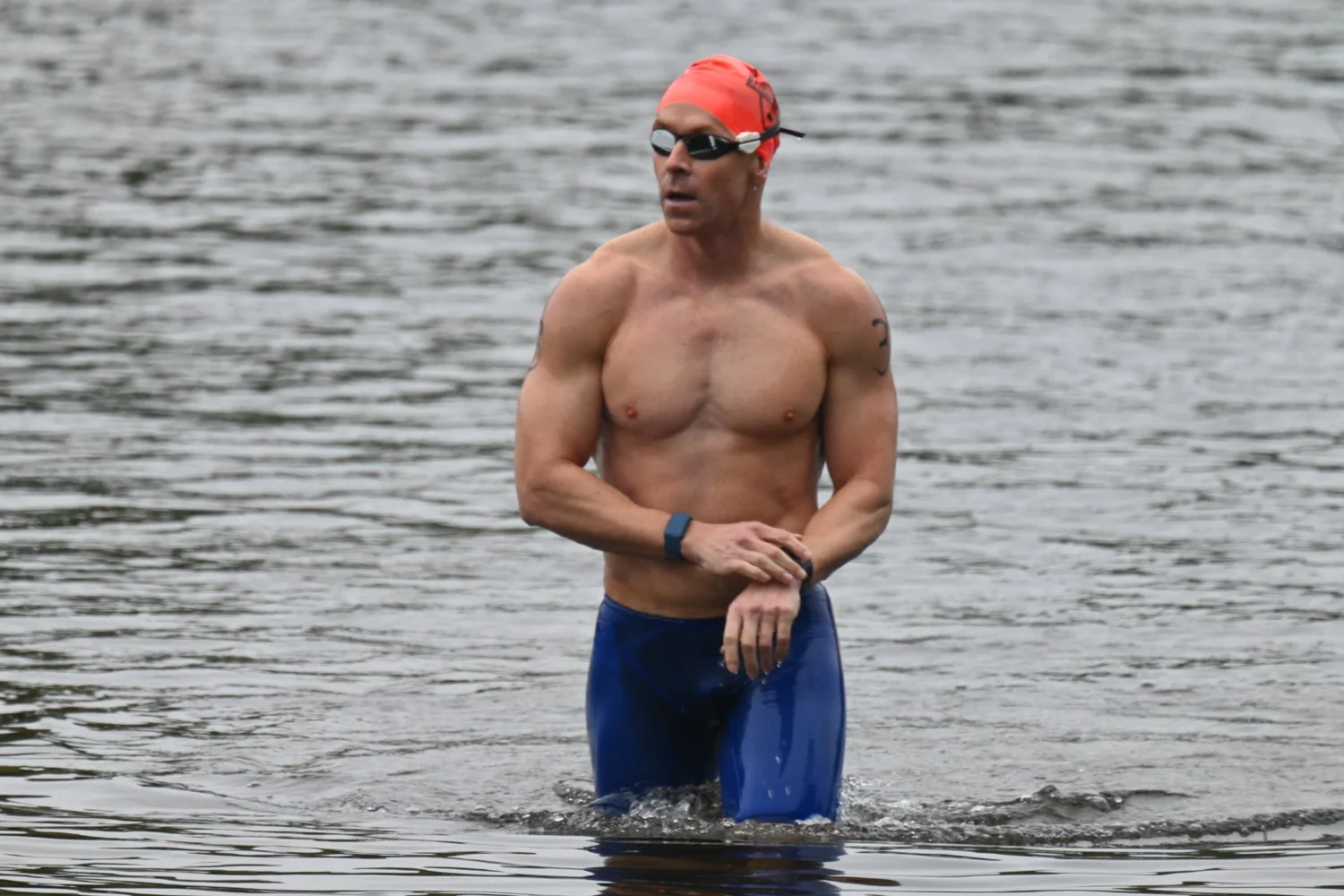 Coach Dan Daly in an orange swim cap and goggles, standing waist-deep in open water.