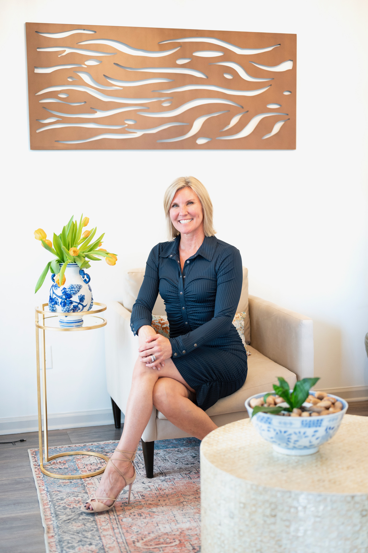 Michelle Johns in a navy dress seated in an armchair in her office with abstract copper wall art behind her