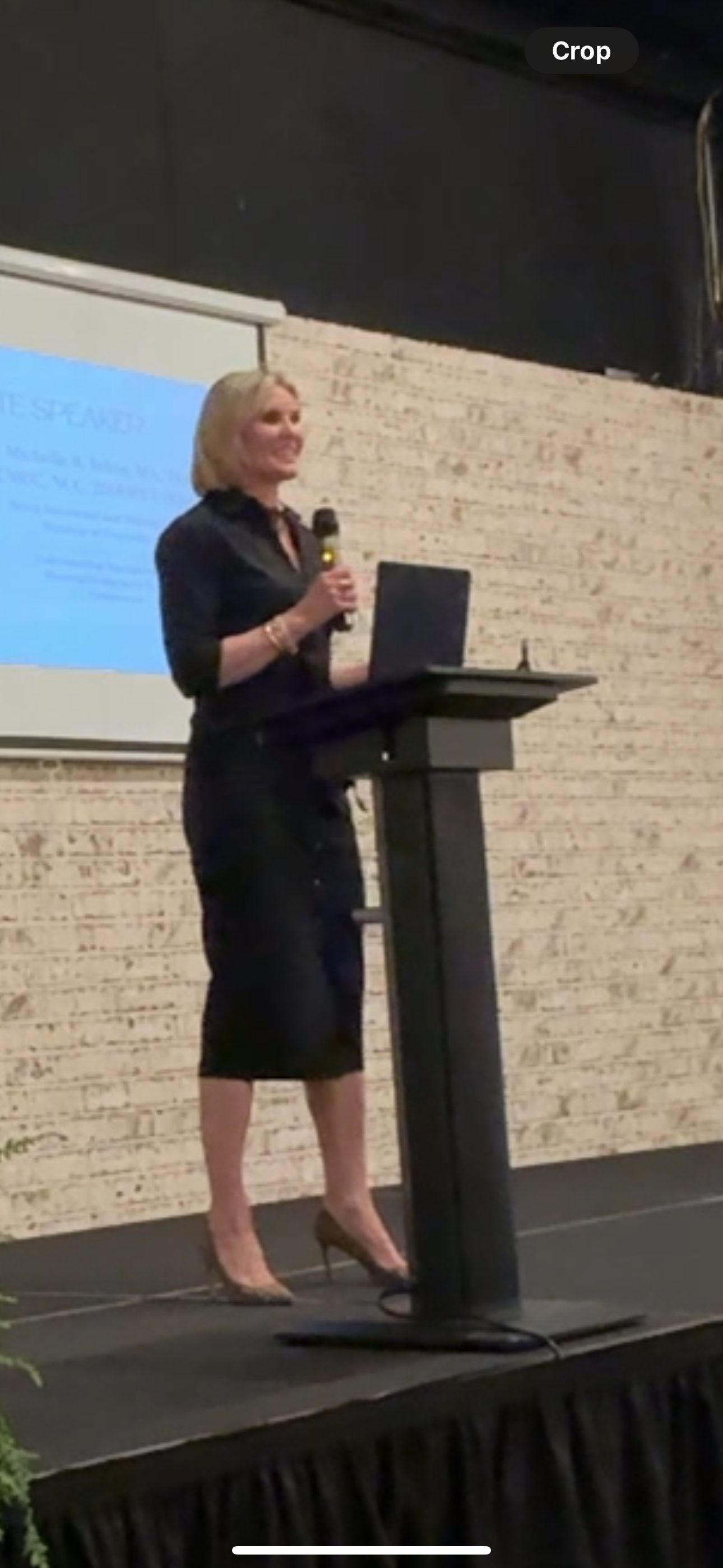 Michelle Johns in a black dress speaking at a podium on stage with a microphone and presentation screen
