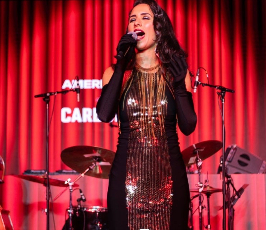 Singer in a gold sequin gown performing before a red curtain