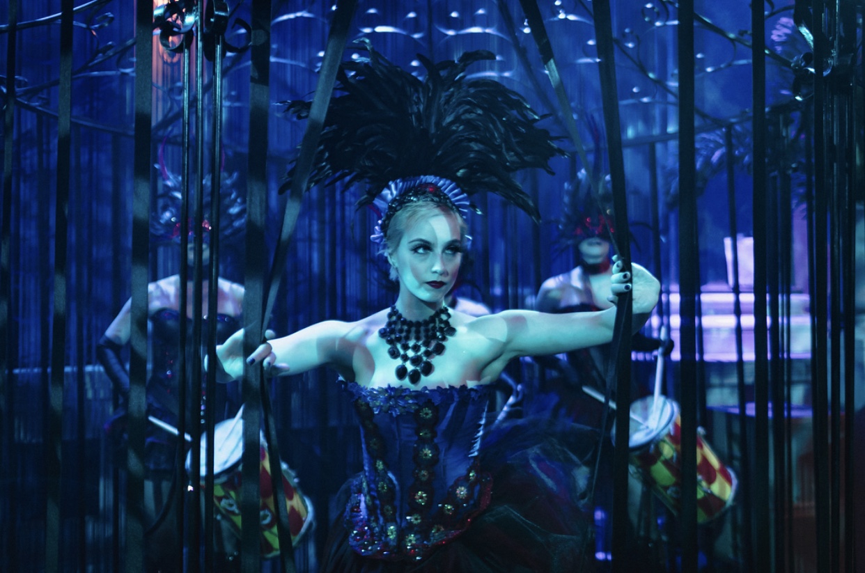 Performer in a blue corset and feather headdress inside a cage-like stage set