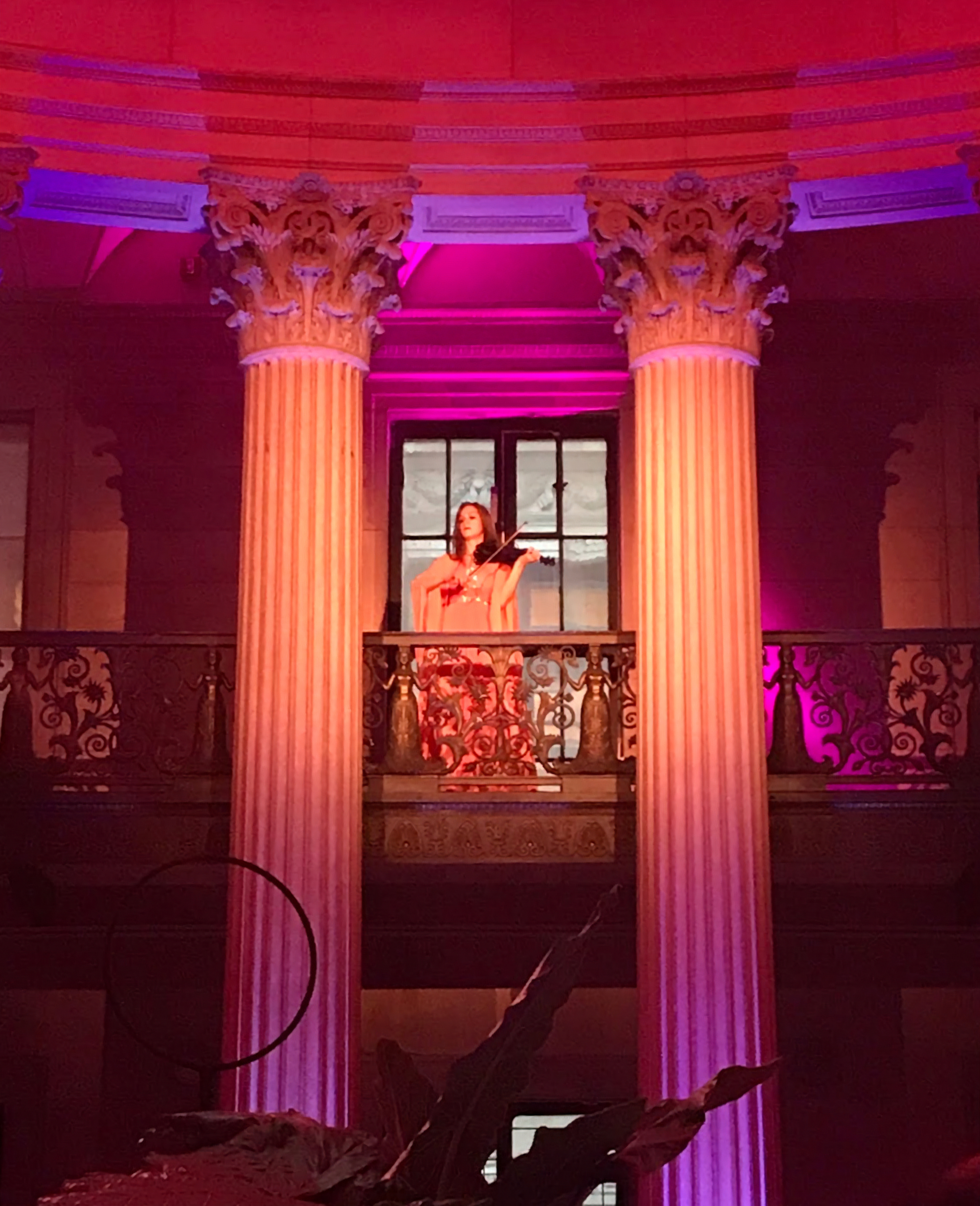 Violinist performing on a grand balcony inside a columned hall with pink uplighting