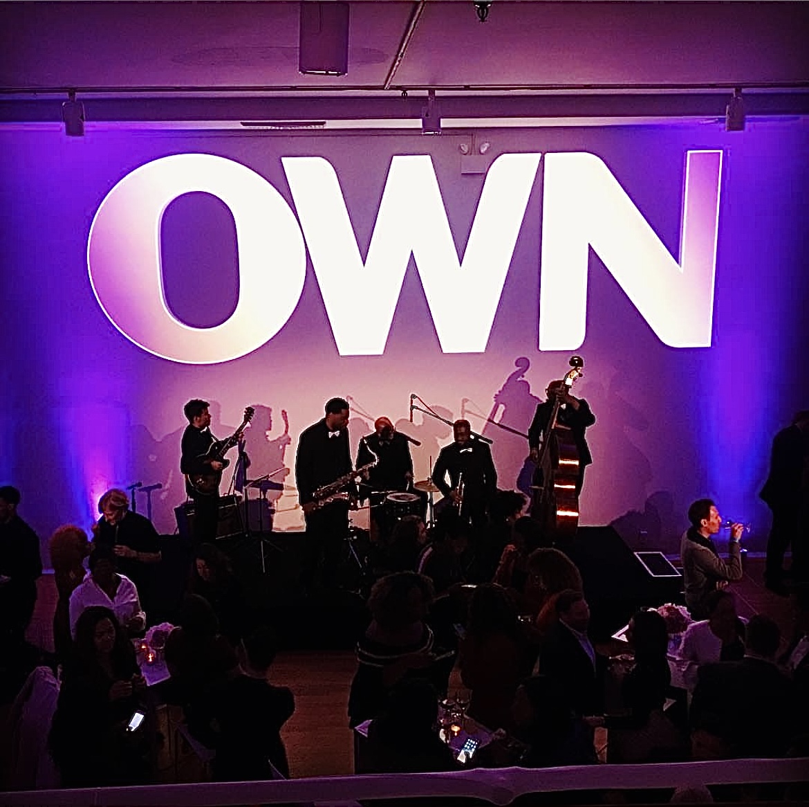 Live jazz band performing beneath a large illuminated OWN logo