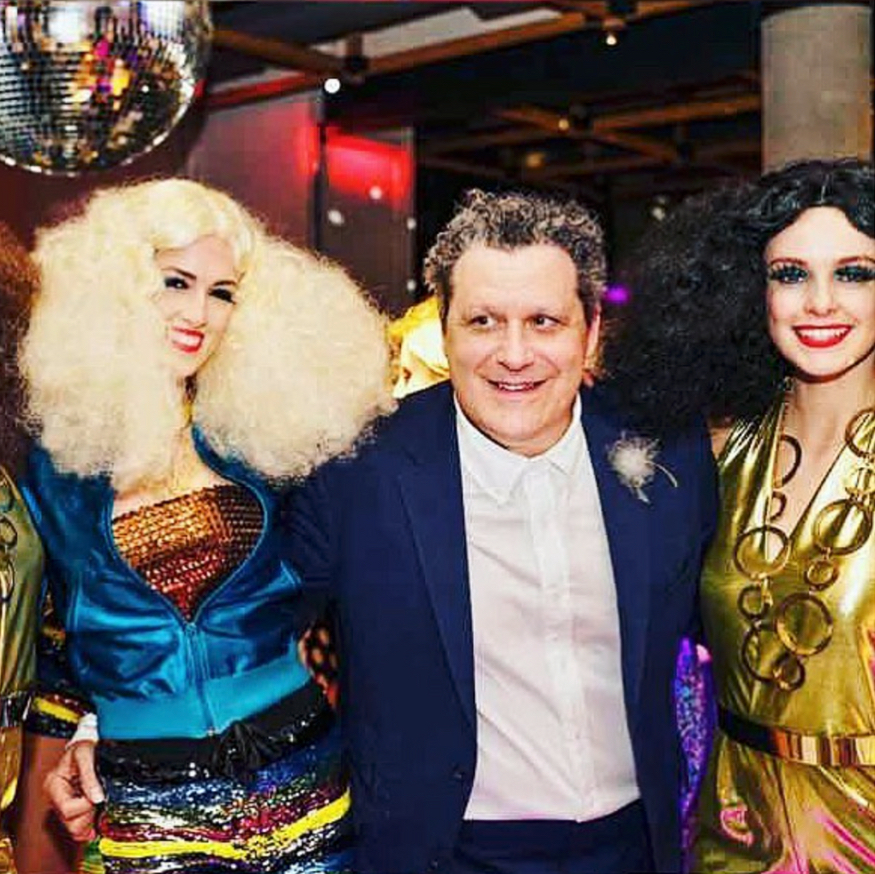 Suited man flanked by two performers in colorful disco wigs and sequins