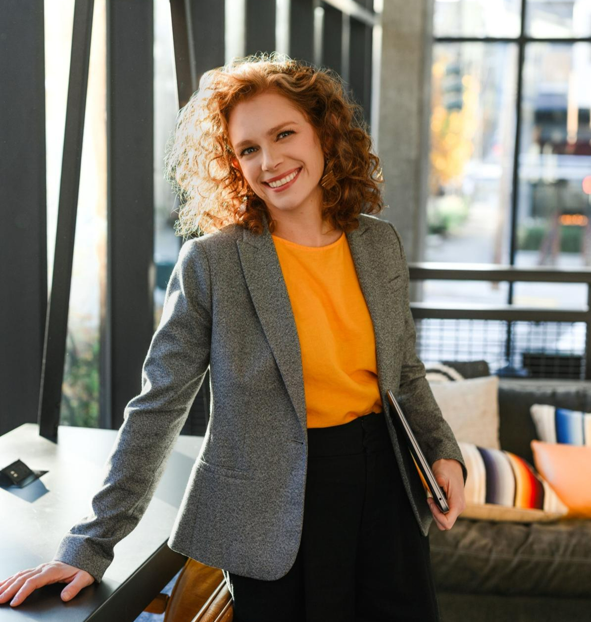 Allie Siegel in a grey blazer and orange top, smiling in an office lobby holding a laptop