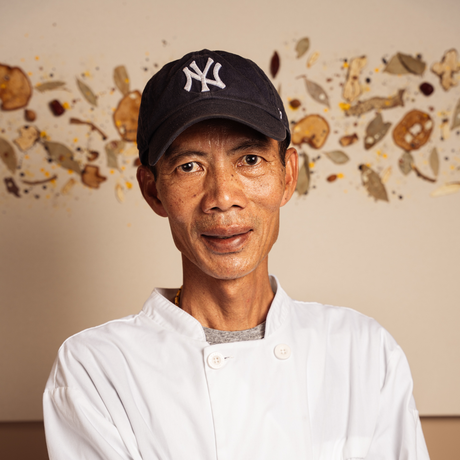 Chef Chen, in a white chef's coat and a navy New York Yankees cap, smiling in front of a decorative spice mural