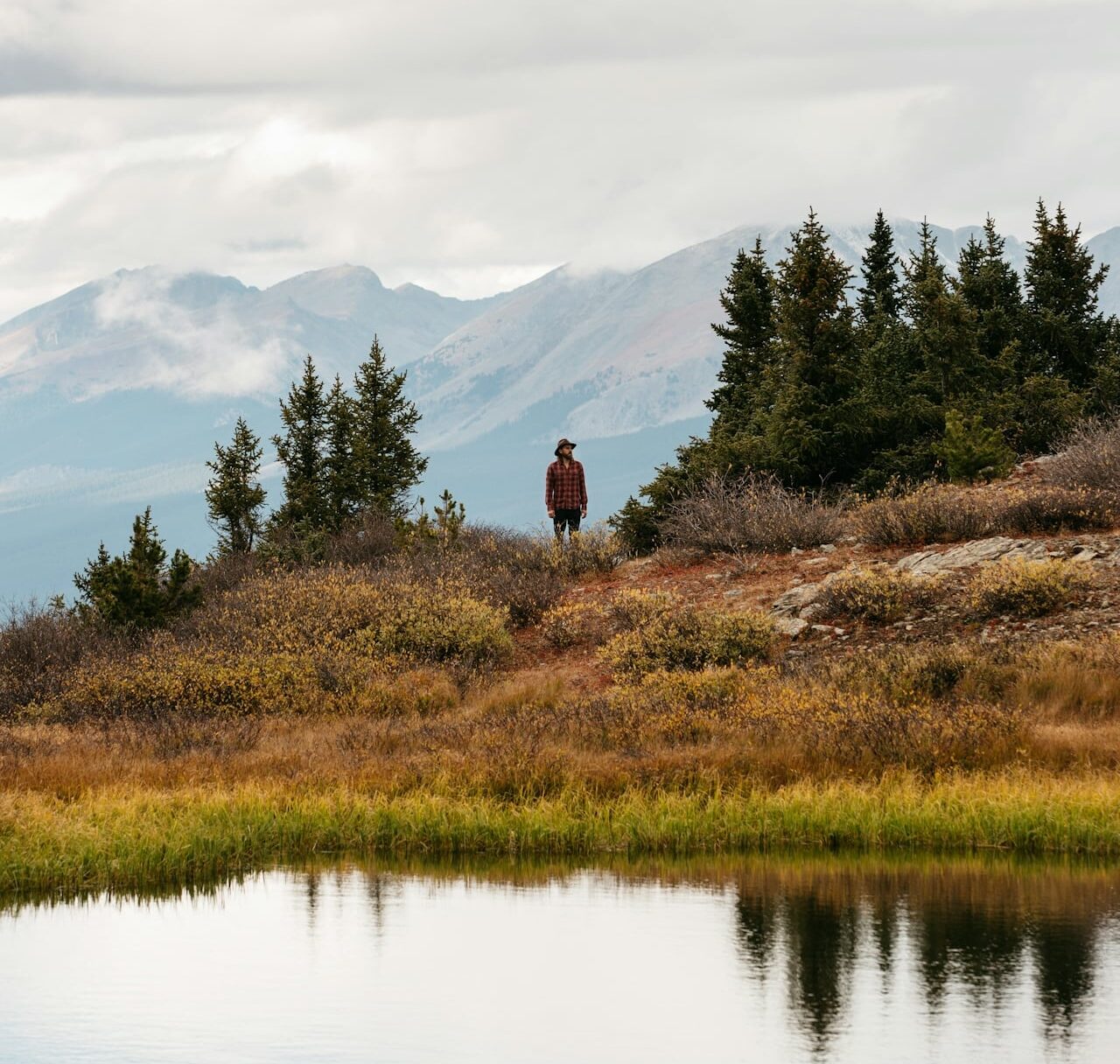 A person in a red plaid shirt standing on a hillside above a mountain lake ringed by evergreens and peaks