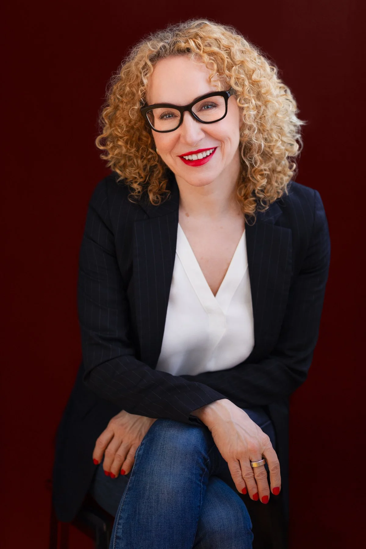 Mila Apostolovic, curly blonde hair, glasses and red lipstick, seated portrait against a burgundy background