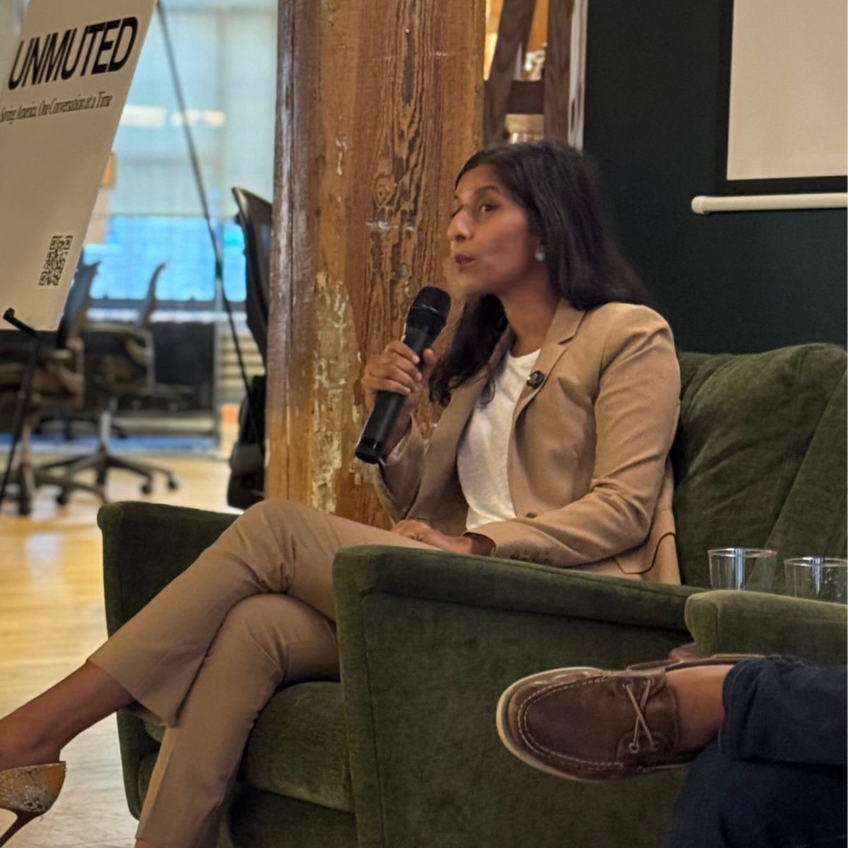 Speaker in a tan blazer holding a microphone in a green armchair at an UNMUTED event.