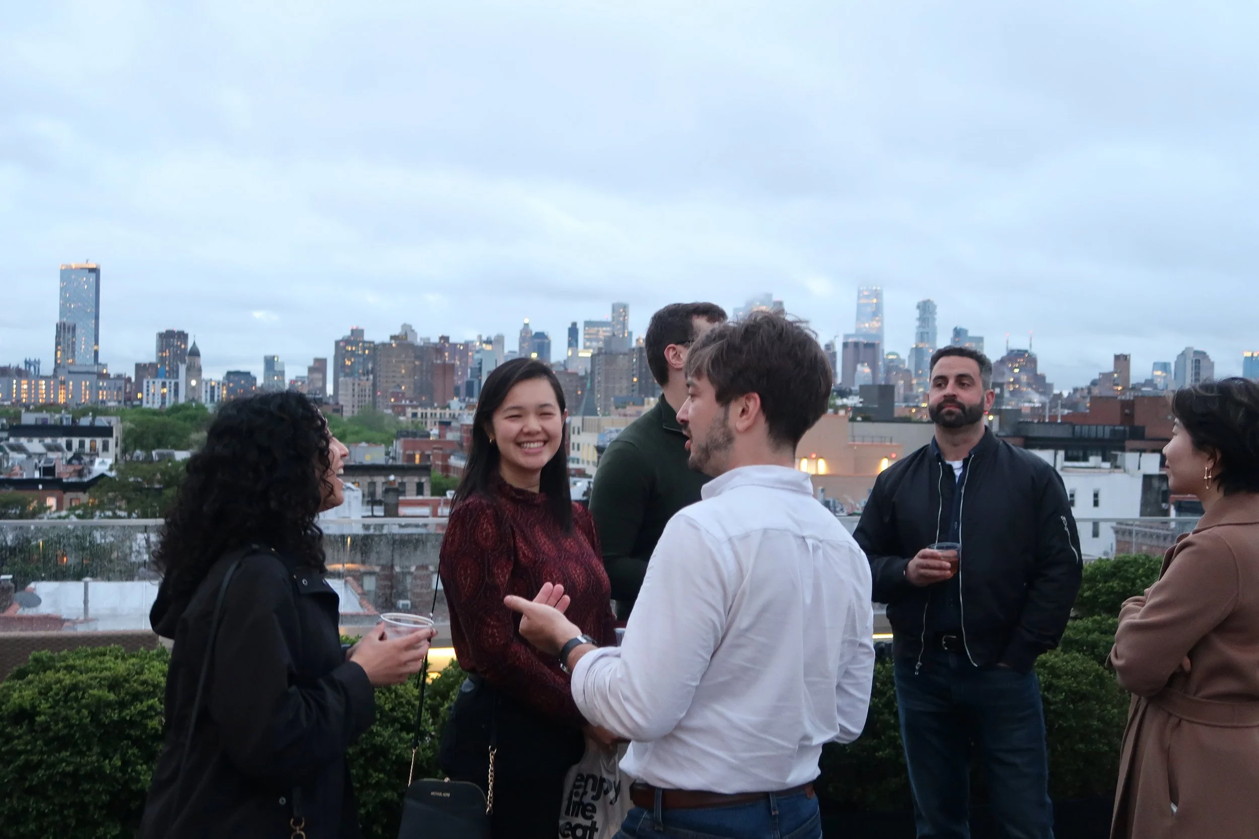 Young adults talking on a rooftop at dusk with the New York City skyline behind them.