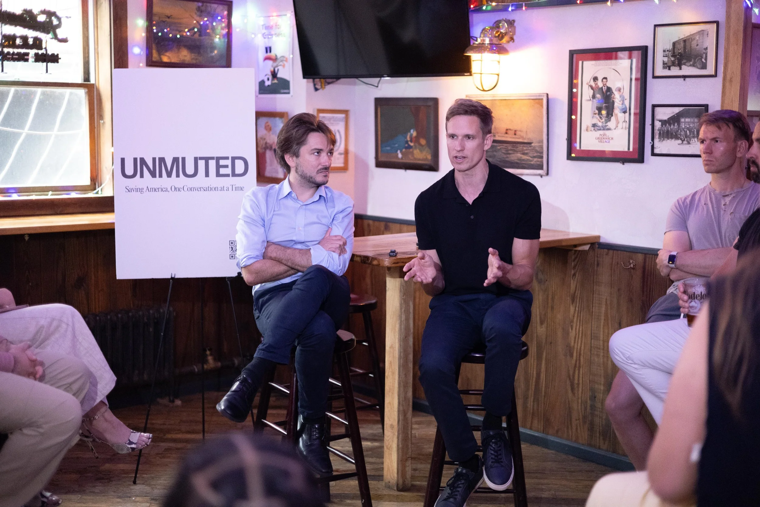 Two speakers on stools at an UNMUTED event beside the UNMUTED sign.