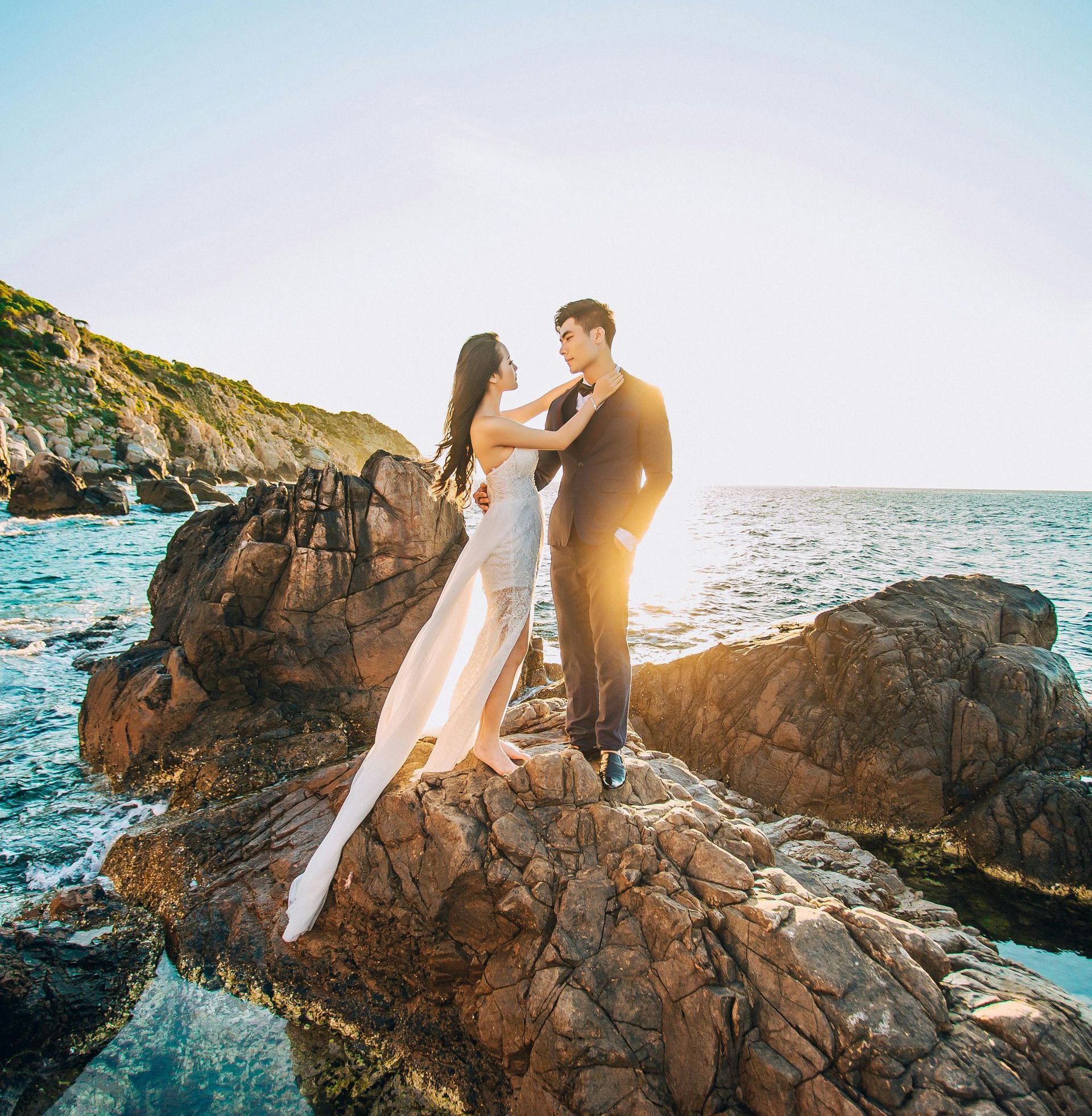 A bride and groom standing on coastal rocks at sunset.