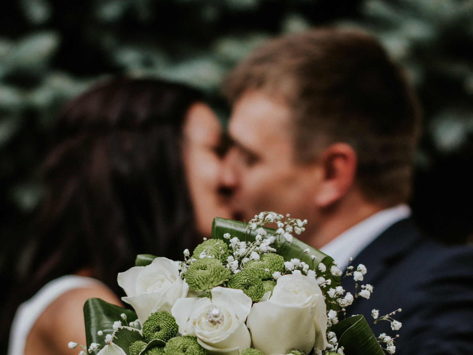 A couple kissing behind a bouquet of white roses and green chrysanthemums.