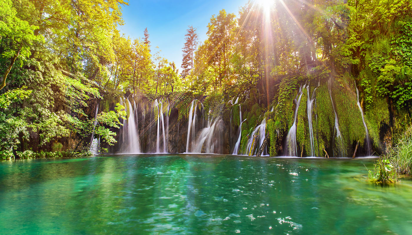 Plitvice Lakes waterfalls cascading into a turquoise pool with sun rays through the forest