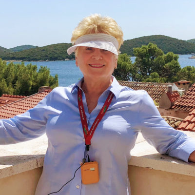 Wanda S. Radetti, blonde woman in a white visor and blue shirt, on a rooftop terrace overlooking the Croatian coastline