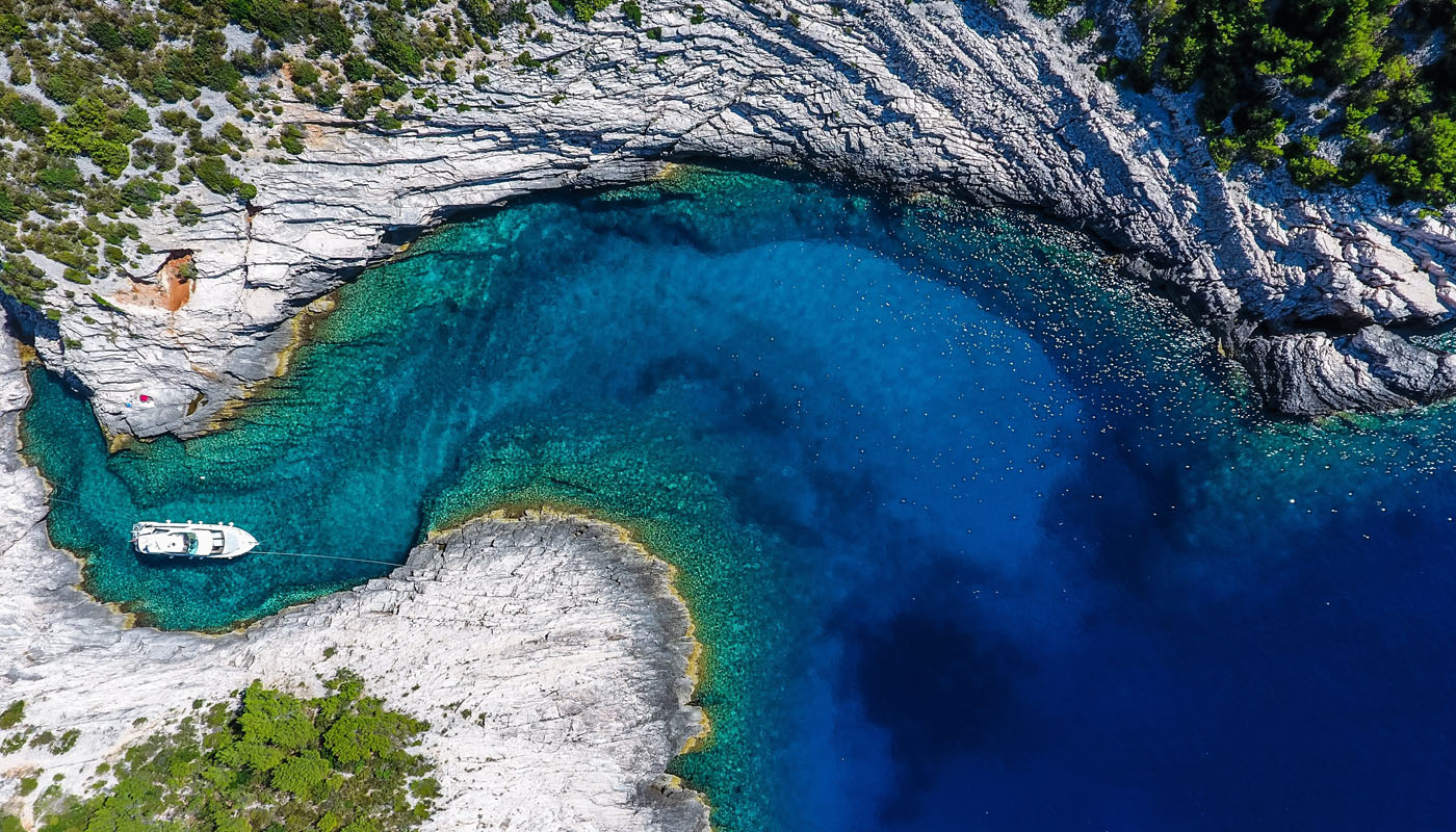 Drone aerial of a rocky Croatian island cove with crystal-clear blue-green water and a single white yacht anchored