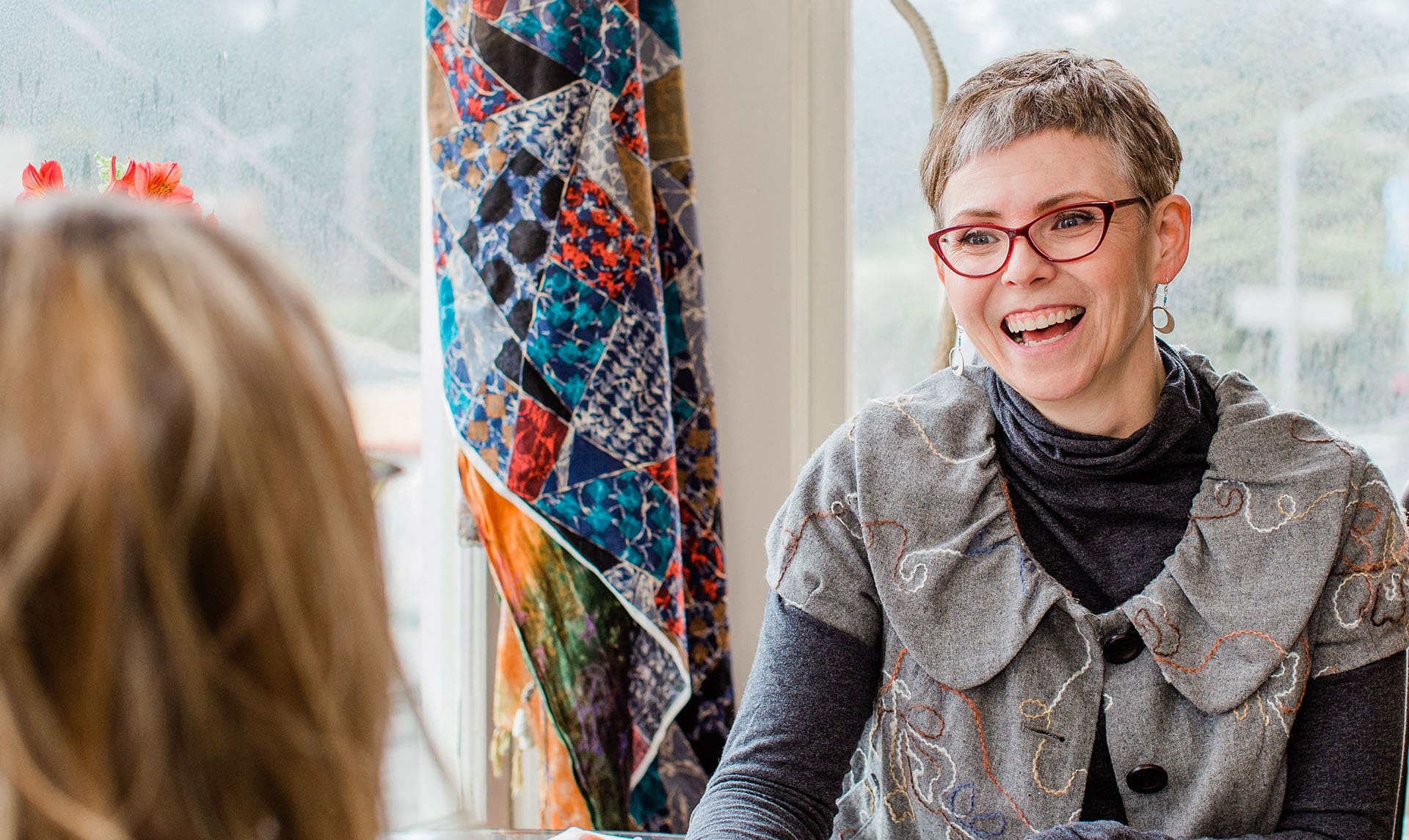 Lauri Smith laughing in red glasses at a table with a colorful quilt behind her
