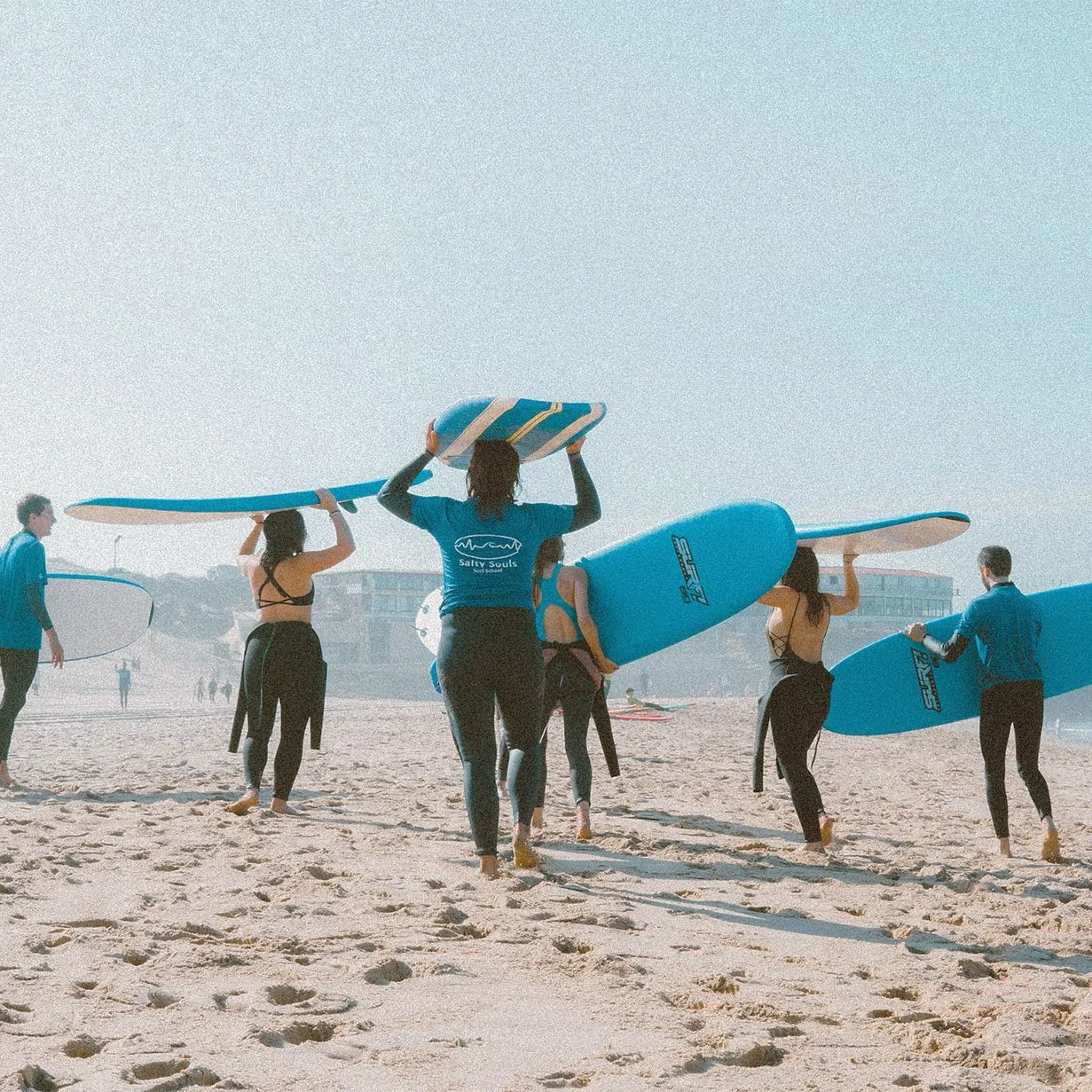 Group of surfers carrying blue boards overhead across the sand on a foggy day