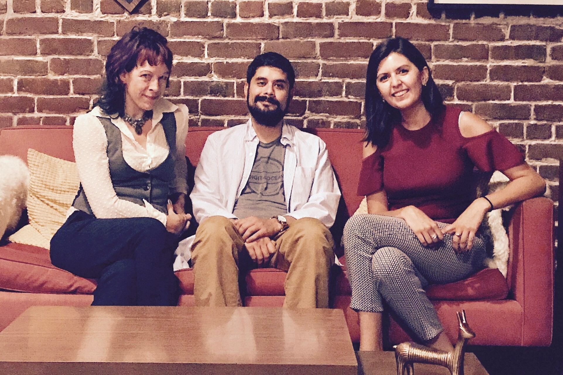 Three panelists seated on a red couch in front of a brick wall hung with clocks and framed prints
