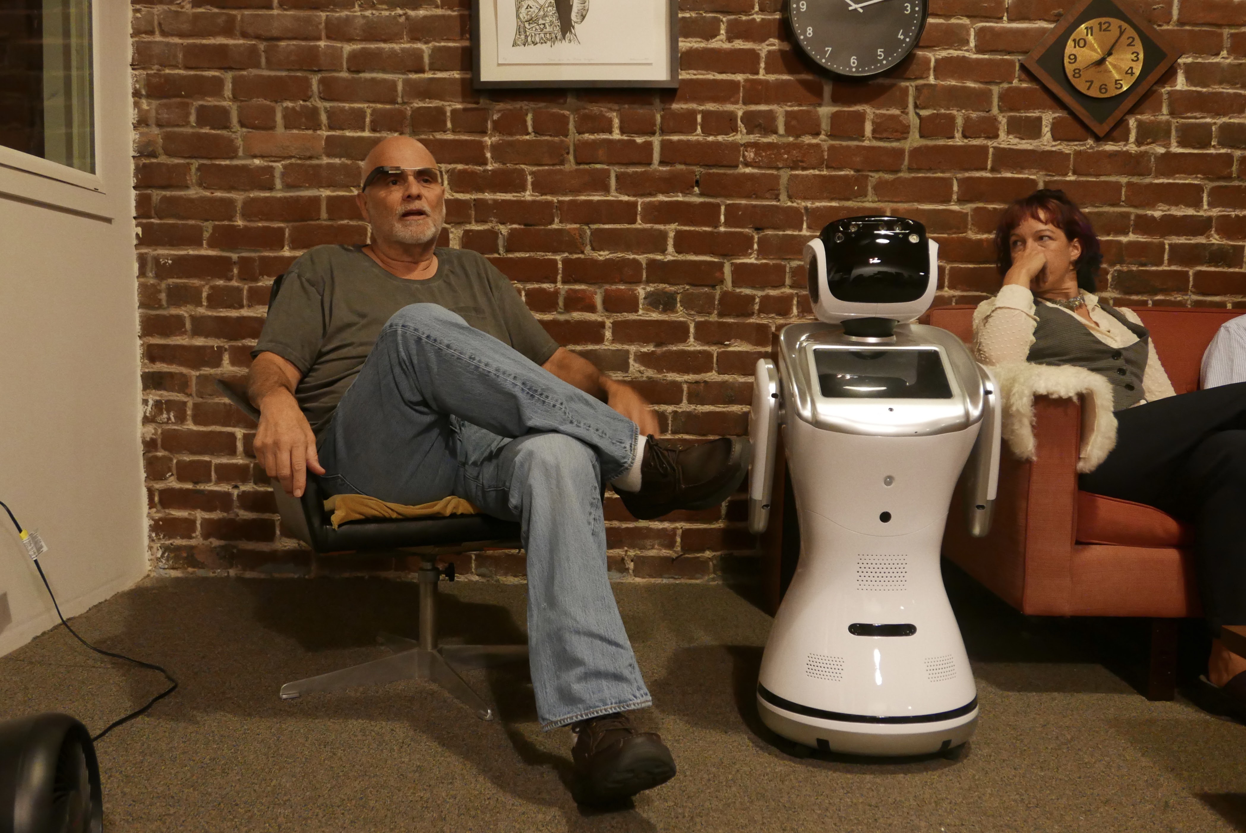 A guest in sunglasses seated beside a Sanbot robot against a brick wall at a WDTnow event