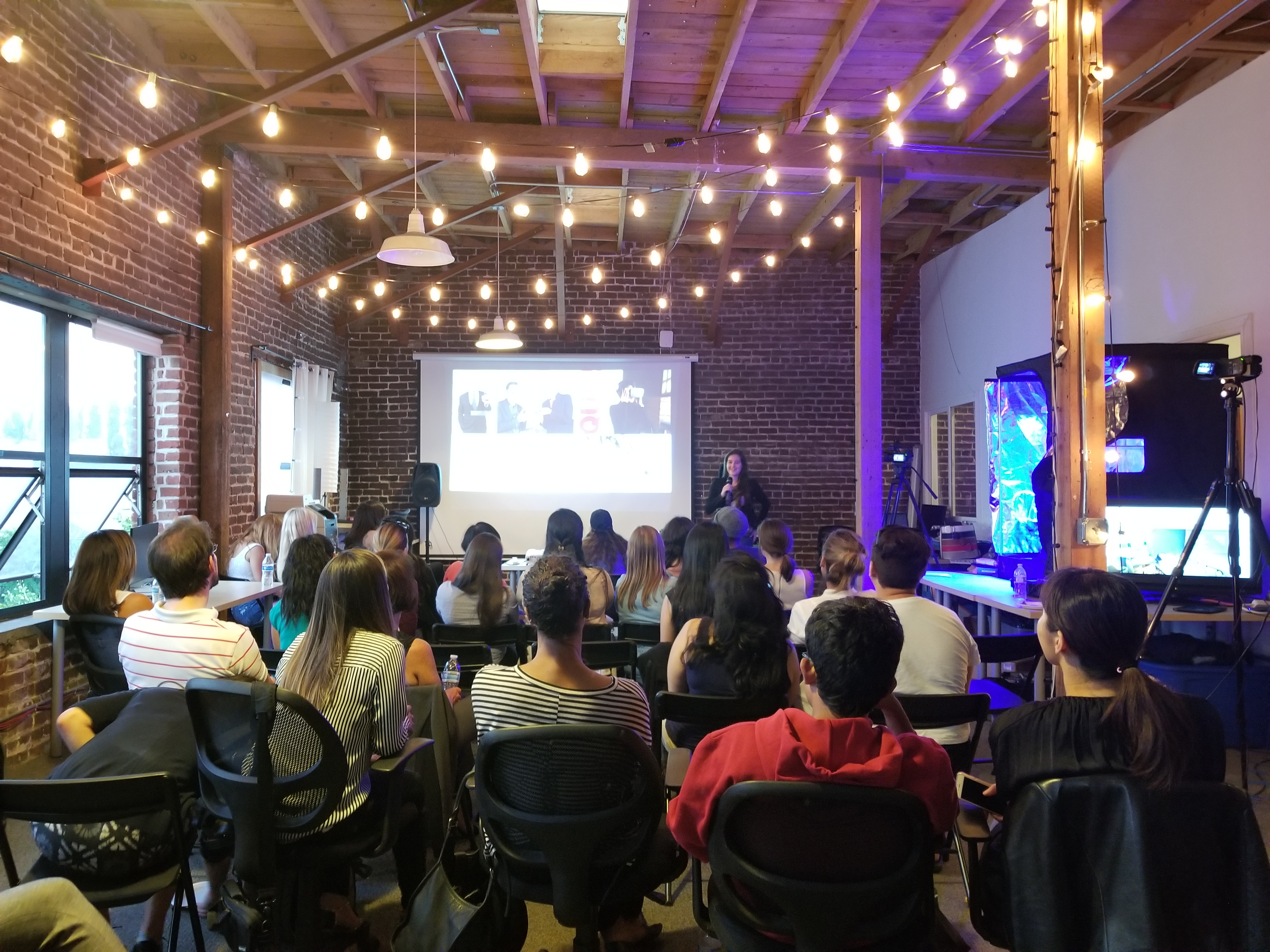 A full audience seated for a talk under string lights, with a speaker and projection screen at the front