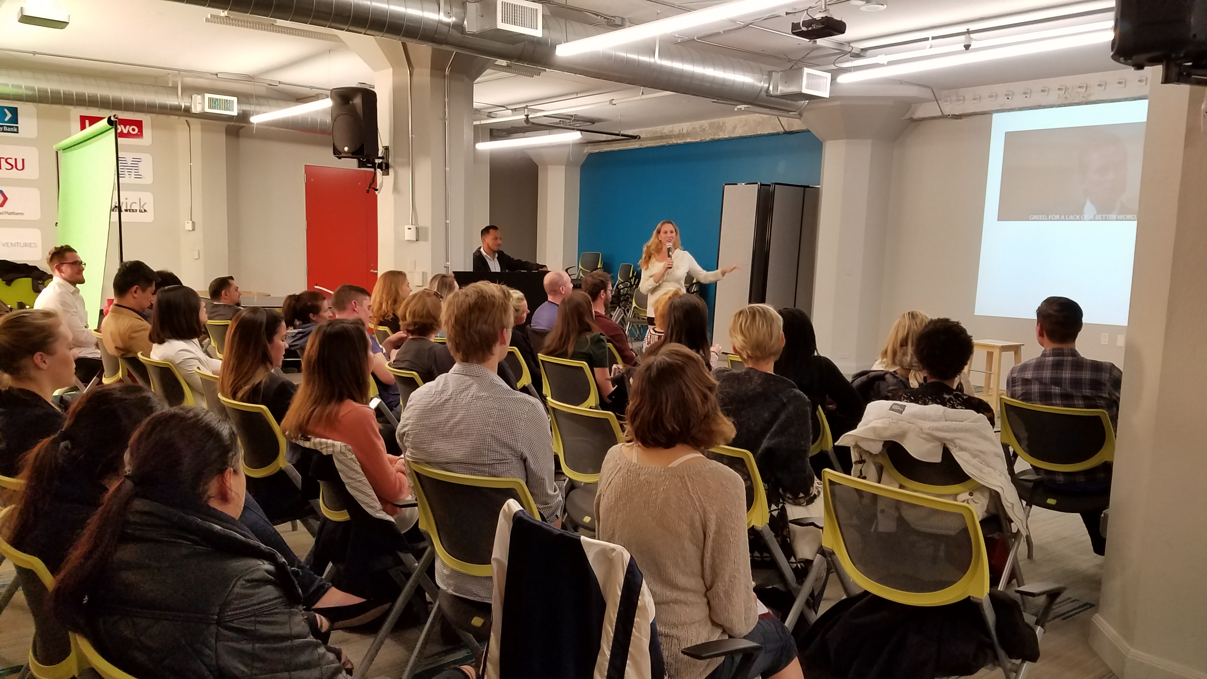 A large audience on yellow-green chairs watching a speaker with sponsor logos on the wall behind