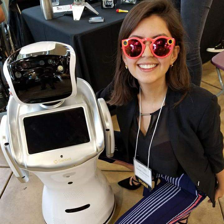 A speaker in red Snapchat Spectacles smiling beside a Sanbot robot at a tech conference