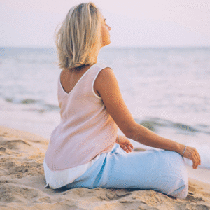 Woman seated on the sand at the beach in a relaxed meditative pose at sunset.