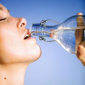 Woman drinking water from a glass bottle against a blue sky.