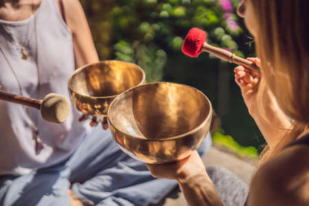 Two women playing golden Tibetan singing bowls outdoors in a garden