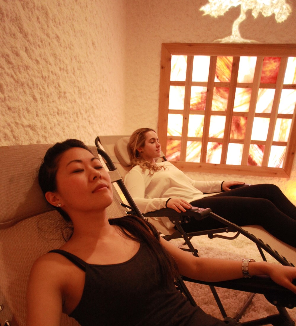 Two women relaxing in reclined chairs inside the Qi Garden Salt Spa beside a glowing Himalayan salt wall