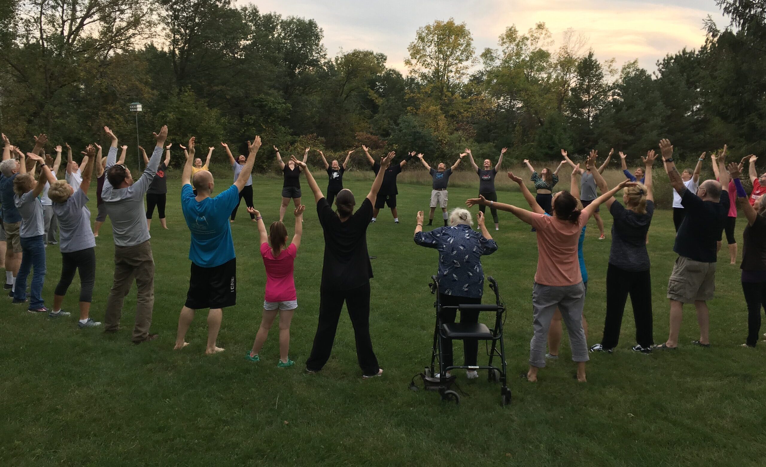 Large group outdoors on a grass field with arms raised during a qigong practice at dusk