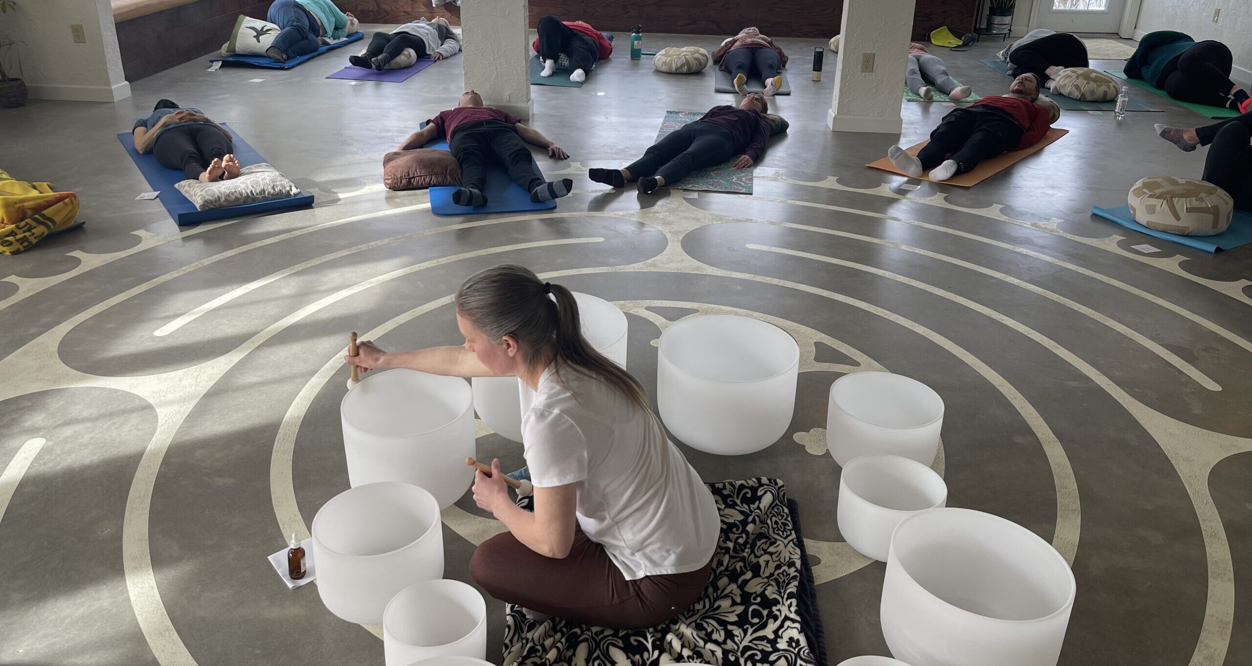 Instructor playing crystal singing bowls in the studio while participants rest on yoga mats over the labyrinth floor