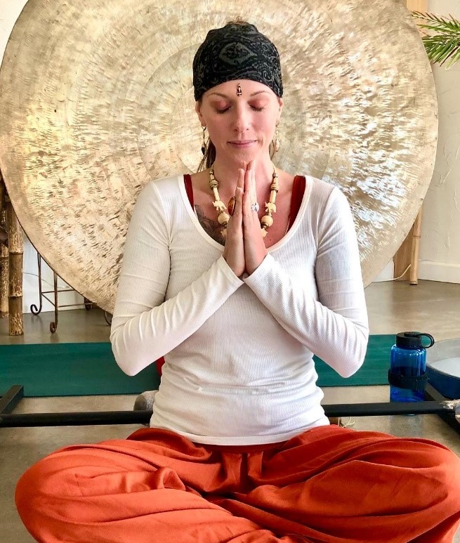 Instructor seated in meditation with hands in prayer in front of a large gong